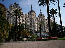 Photographie en couleur d'une grande façade claire et ouvragée sur fond de ciel bleu, avec des palmiers