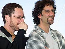 Photo de deux hommes blancs souriants s'arrêtant à la poitrine. Celui de droite porte des lunettes et une courte barbe, celui de gauche a les cheveux plus longs.