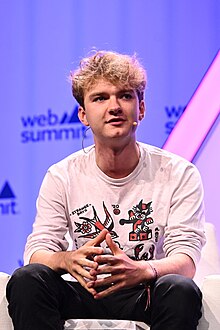 Young man with curly hair dressed casually in multi coloured tee shirt gyrating on a theatre stage addressing the audience by way of a headset microphone