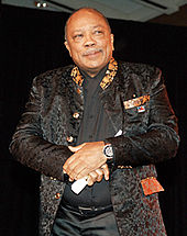Quincy Jones, a plump bald African American man with a grey mustache and wry smile. He is elegantly dressed in a black brocade jacket with a patterned collar over a black shirt.