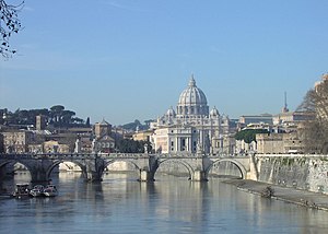 A view of Rome on a sunny afternoon looking along the river. A bridge crosses the river and beyond it is a hill on which the grey dome of St Peter's rises above ancient buildings and dark pine trees.