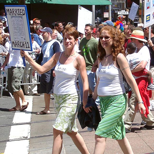 Zu sehen sind zwei Frauen die auf einer Straße in einer Menschenmenge Händchen halten. Beide tragen Röcke und ärmellose weiße Oberteile. Eine der Frauen links im Bild hat kurzes Haar, die andere langes. Die Kurzhaarige hält ein Schild hoch uf dem schlecht erkennbar die Worte „Married...“ zu lesen sind.