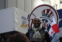 Мариано Ривера в темном полупальто и сером шарфе улыбается, держа в руках красно-бело-синий флаг. Он стоит перед красно-бело-синим логотипом с надписью «Янки».
