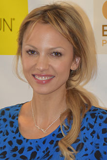 A blonde woman wearing a blue and black shirt smiles towards the camera.