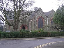 A Victorian church in stone with brick detail and a slate roof, separated from an urban road by a hedge. There are large arched windows.