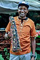 Mann med tradisjonelt musikkinstrument i ein pose pynta med perler, iført ei skjorte med perledekorasjonar.