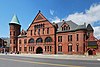 An ornate and complex dark red brick building, about three and a half stories tall, seen from across a wide street. It has a gabled front section, tall green-roofed conical tower on the left, and a lower engaged corner tower on the right.