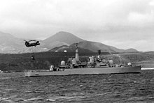 A twin-rotor helicopter with a load slung below hovers over a warship