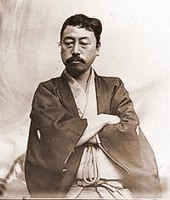 Портрет азиатского мужчины с усами, одетого в традиционную японскую одежду. Он смотрит вниз, скрестив руки.