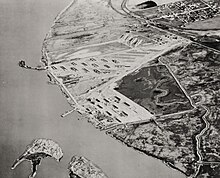 Aerial photograph, looking eastward, taken between 1942 and 1944. The town of Port Chicago is in the upper right. The lower left shows utility and personnel piers extending toward the two sections of Seal Island. The munitions loading pier curves to the left beyond 20-odd revetments. Marshy tidal zones separate the munitions pier from barracks buildings near the personnel pier and near the town. PortChicago-aerial-construction.jpg