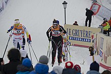 Deux skieuses de fond de face dans une montée, avec au premier plan des têtes de spectateurs, de dos.