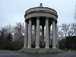 Open air round, marble structure resting on a layered base with twelve columns supporting the round, marble roof. There are leafless trees in the background