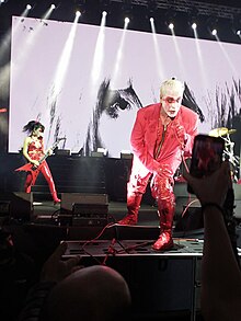 Lindemann trägt rote Kleidung und blondierte Haare, hat ein rot-weiß geschminktes Gesicht und hält nach vorn gebeugt ein Mikrofon in der Hand. Im Hintergrund ist eine Musikerin mit E-Bass zu sehen.