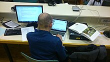Un jeune homme assis devant un ordinateur de l'espace numérique de la Part-Dieu avec son propre ordinateur portable et un livre de la bibliothèque en train d'enrichir une fiche Wikipédia.