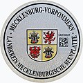 Aktuelle Zulassungsplakette des Landkreises Mecklenburgische Seenplatte mit dem mecklenburg-vorpommerschen Landeswappen, rechts mit DataMatrix-Code und Landkreiswappen oben links