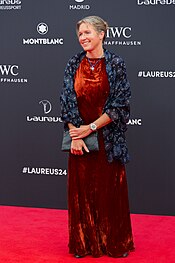 The image shows Kirsten Neuschäfer on the red carpet at the 25th Laureus World Sports Awards. The person is elegantly dressed in a floor-length red velvet dress and a dark blue patterned jacket, holding a grey clutch. She stands in front of a backdrop featuring the logos of Montblanc, IWC Schaffhausen, and the hashtag #LAUREUS24.The overall ambiance is one of luxury and celebration of sporting excellence.
