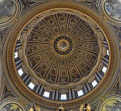 Photo looking up at the dome's interior from below. The dome is decorated at the top with a band of script. Around its base are windows through which the light streams. The decoration is divided by many vertical ribs which are ornamented with golden stars.