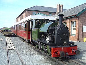 A green saddle-tank engine stands in front of a large brick station building. Behind the engine are four red coaches.