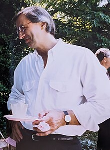 Jimmy is a tall man witth short gray hair. He's holding a small party plate and smiling toward someone off-camera.