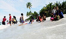 A photo of dark-skinned boys and girls standing, sitting and lying in wet clothes on the surf of a beach. Some palm trees stand in the background. Some women in veils are among the kids.