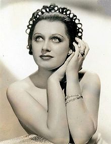 A young white woman with an elaborate looped hairstyle, bare shoulders and arms clasped before her, in a glamor-style publicity photo