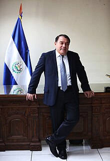 A full-body portrait of Reinaldo Carballo leaning on a wooden desk and facing the camera