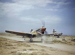 Kittyhawk Mk.III 112. peruti při pojíždění v Medeníně v Tunisku, 1943. Příslušník pozemního personálu sedící na křídle navádí pilota jehož výhled vpřed je omezen předkem letadla.