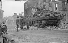 Photographie montrant un char de combat dont sortent deux soldats au bord d'une rue en ruine et quatre autres soldats attendant au centre de la rue