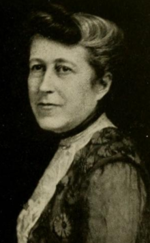 An older white woman, her grey hair in an updo, wearing a high-collared lace blouse or dress