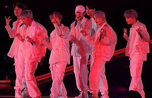 Seven men dressed in white performing on stage illuminated with pink lights against a black backdrop