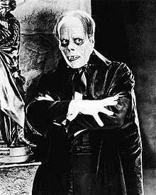 A man wearing extensive makeup is looking to his left. The makeup gives a skull-like appearance and he is wearing a robe with his arms crossed in front of him.