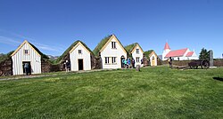 Several turf houses in a row with a broad field of grass in front of them. There is a church a little ways from the houses.