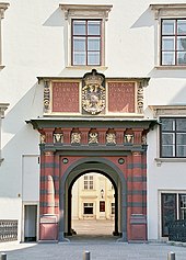 Frontale Farbfotografie von einem Durchgang in grün-braunen Querstreifen mit goldenen Ornamenten, der in die weisse Fassade mit Fenstern eingebaut ist. Über dem Durchgang befinden sich ein Wappen und lateinische Wörter.