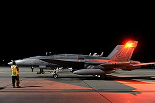 Side view of military combat jet with external fuel tanks and wingtip missiles, parked on airfield at night with ground crewman in foreground