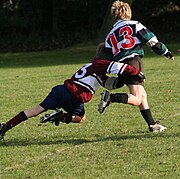 A child running away from camera in green and black hooped rugby jersey is in the process of being tackled around the hips and legs by an opponent