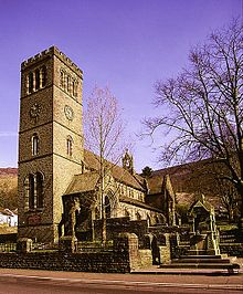 A stone church with a tall square tower, in the foreground is a stone wall and fountain