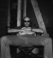 A black and white photo of Ted Stamm in jeans and dark sunglasses slouched in a director's chair. Stamm has his legs spread far apart and his hands on his chest.