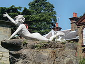 Цветная фотография отдельной женской статуи с короткими взъерошенными волосами, лежащей на каменном постаменте. Опираясь на левую руку, ее голова поднята, а правая рука поднята.