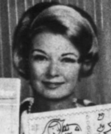 A smiling white woman with a blond bouffant hairdo, holding pictures of stamps