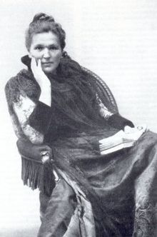 A photograph of a woman seated in a chair with one hand on her right cheek and the other holding a book in her lap