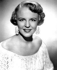 A black-and-white photograph of a woman with short hair and a white shirt. She is looking toward the camera.