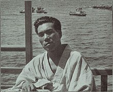 Teikō Shiotani, when 35 years old. He is wearing a yukata and faces the camera across a table. The sea is visible behind him.