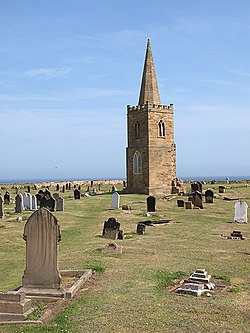 A two-stage church tower, topped with a steeple, surrounded by gravestones, and a view of the sea beyond