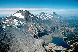 Aerial view from the southeast looking north of the Three Sisters volcanoes, three mountains sparsely covered with ice and snow. From left to right the image shows South Sister, Middle Sister, and North Sister, with a black lava flow in the left foreground.