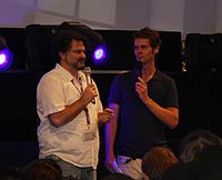 Two men, one with dark hair and older, the other younger with brown hair, speaking in front of an audience.