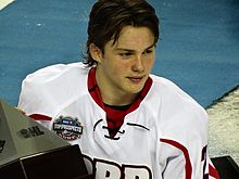 Photographie couleur d’un homme, jeune, vêtu d’un maillot de hockey sur glace blanc, transpirant, le regard porté sur sa gauche