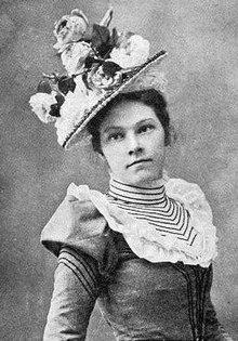 A young white woman wearing a hat covered in large flowers, a dress with a striped high collar and ruffled yoke.