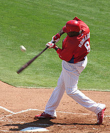 A left-handed man in a red baseball jersey and white baseball pants with red pinstripes swings a black baseball bat at a baseball. His jersey reads "Howard" and "6" on the back in white print.