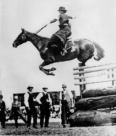 A woman riding sidesaddle clearing a high competition jump in a showground.  There are men beside the jump watching her in admiration of her skill.
