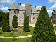 Le château de Cordès est photographié derrière un parc à la française avec allée en terre battue, bassin circulaire en eau, conifères taillés en pointe. Façade en pierres noires apparentes et toitures grises. Le ciel bleu est clairsemé de nuages blancs. Sur la droite de la photo, un bâtiment annexe avec une table.